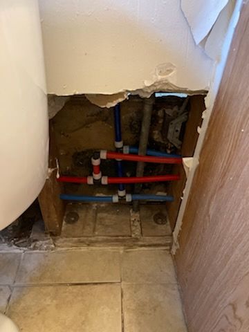 Pipes exposed behind damaged wall in a bathroom, with red and blue PEX tubing visible.