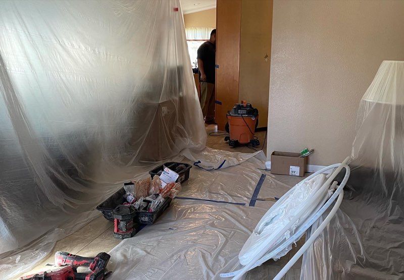 Room under construction, plastic sheeting protecting furniture. Person in doorway, equipment, tools and materials on the floor.