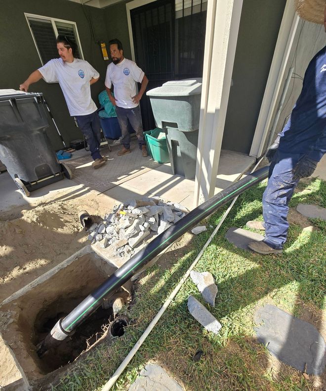 Workers installing a black pipe in a trench next to a building. Two watch nearby.