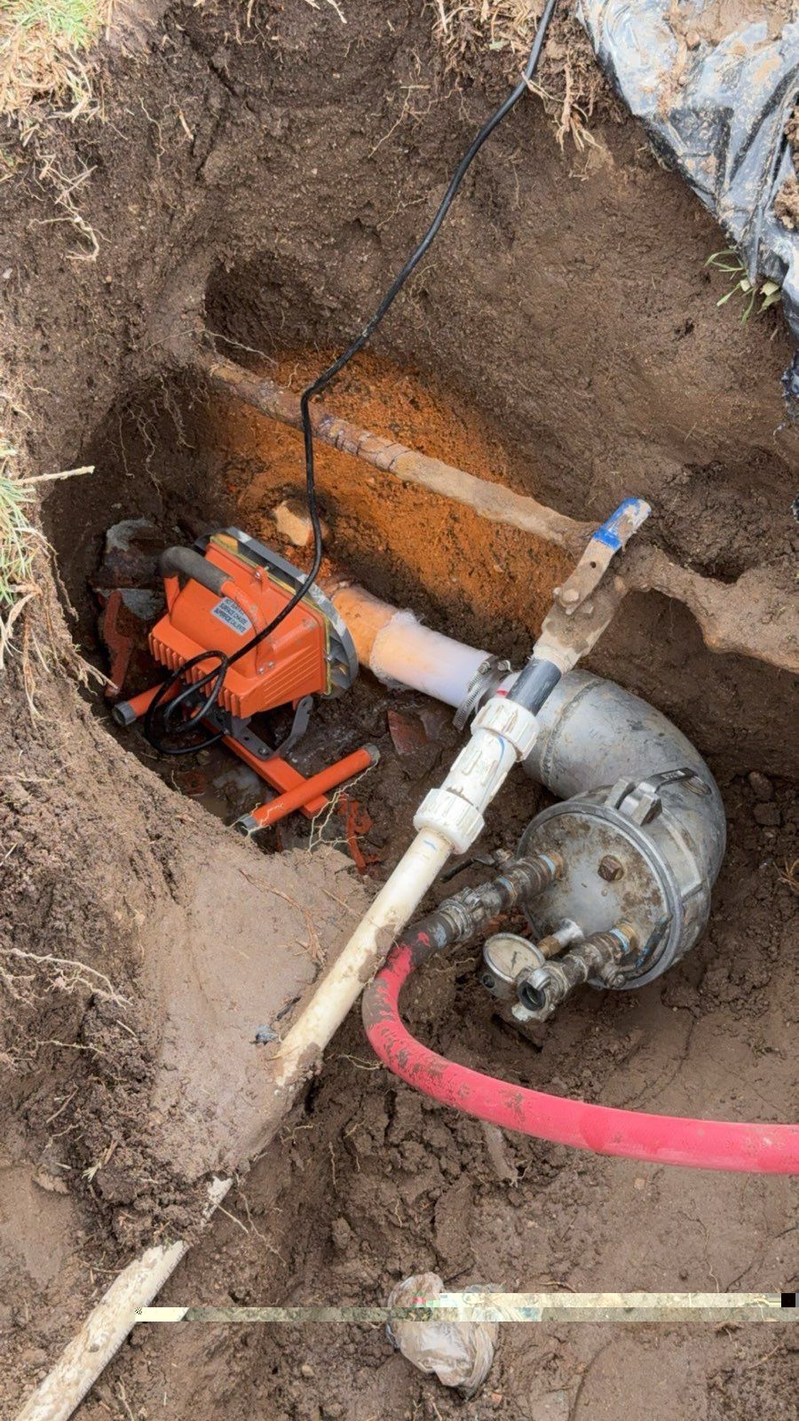 A chainsaw and pipes exposed in a dirt pit. An orange saw lies next to the metallic plumbing.