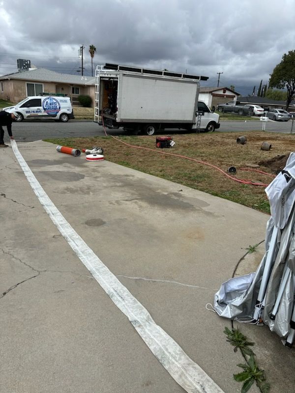 A white truck is parked on a lawn. A hose lies on a driveway. A person tends to the hose. Cloudy sky.