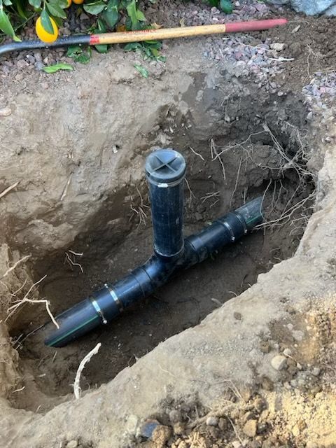 A sprinkler system's elbow-shaped pipe fitting buried in a dirt pit. Black pipe and fitting with a sprinkler head.