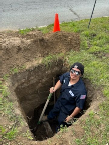 A person in a blue uniform and sunglasses inside a deep hole, holding a shovel. Orange cone on the ground.