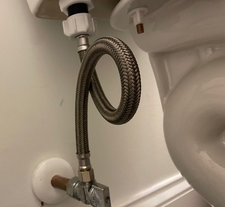 A flexible, braided water supply line coiled between a toilet and a shut-off valve on a white wall.
