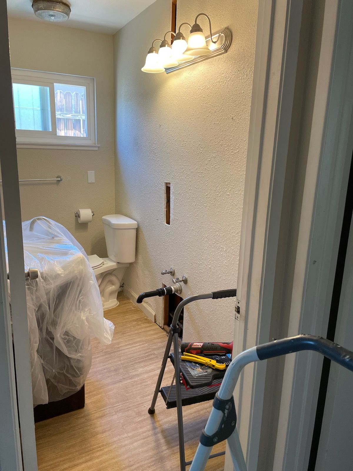 Bathroom renovation in progress; toilet, vanity covered, tools on a step stool, exposed wall.