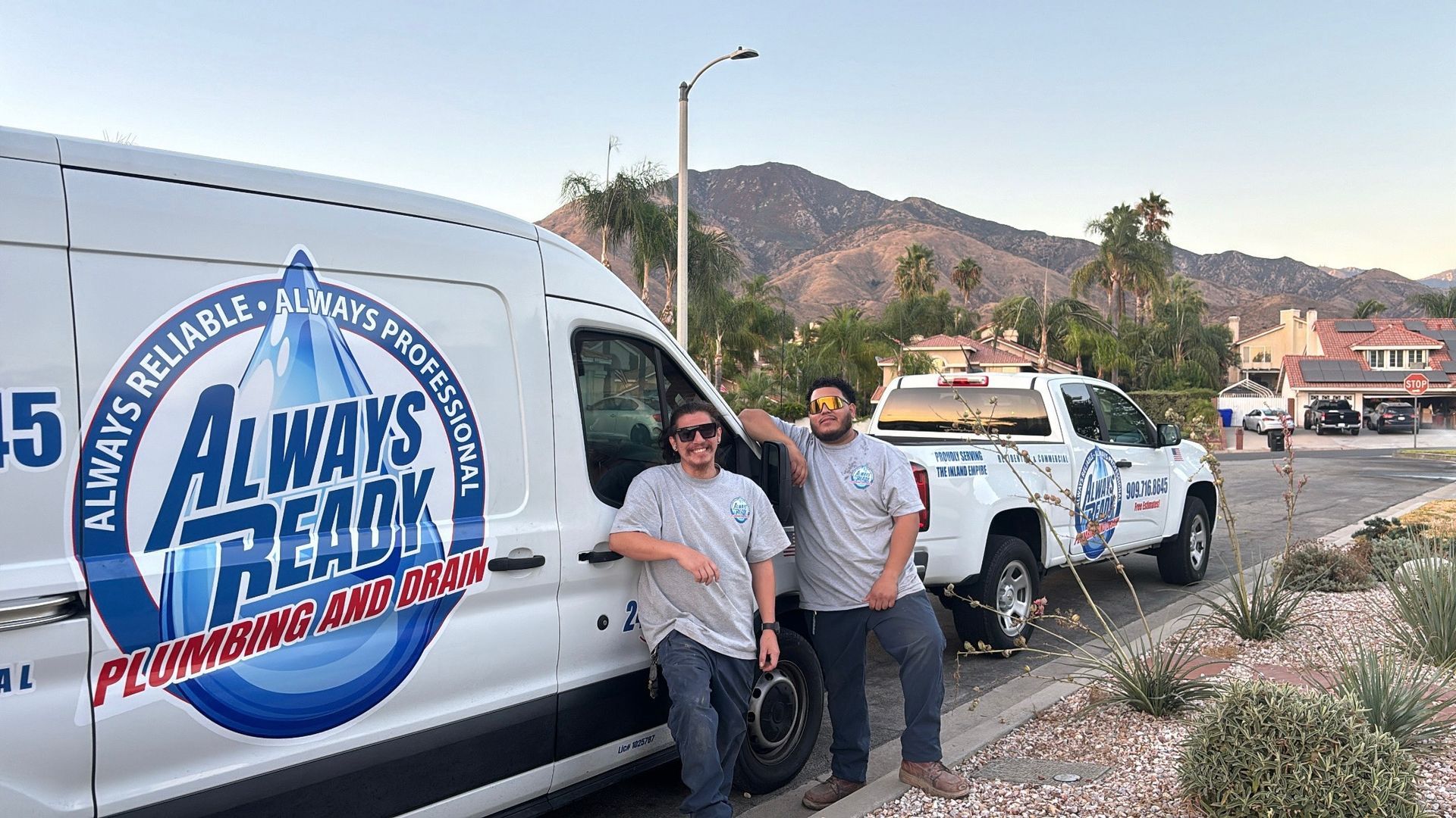 Two men stand by plumbing service vehicles with a mountain backdrop. 