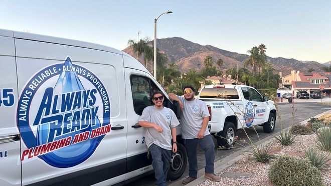 Two men stand by plumbing service vehicles with a mountain backdrop. 