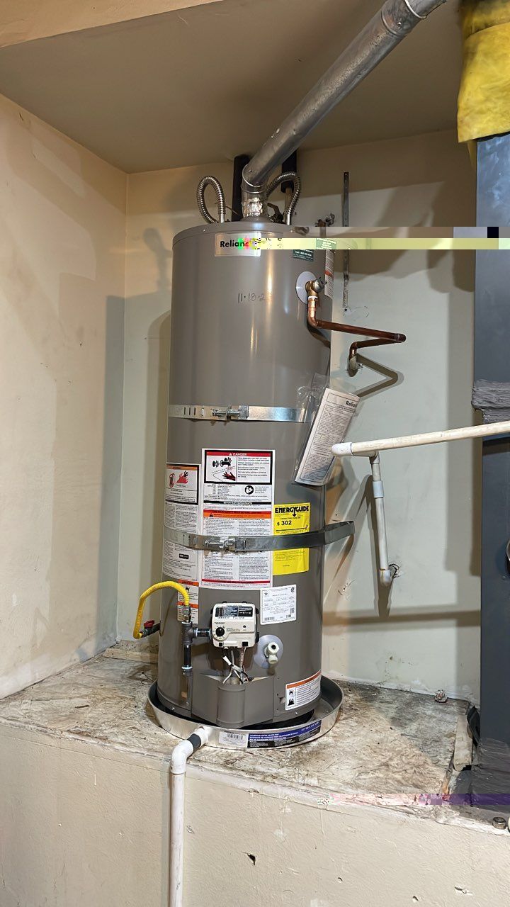 A tall, cylindrical water heater in a basement. It's gray and has various labels and pipes attached.