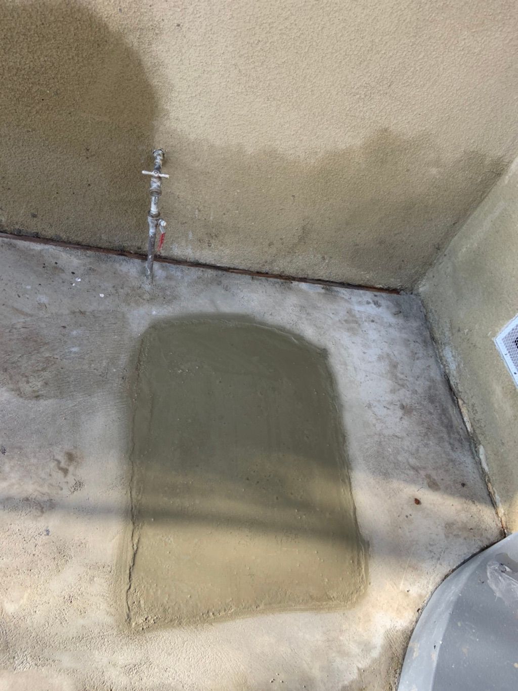 Patch of wet cement on a light-colored concrete surface near a water pipe on a wall.