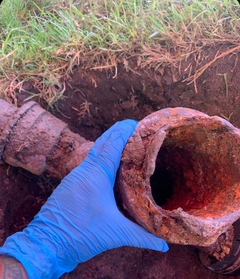 A gloved hand holds a rusted pipe segment, unearthed from soil, near grass.