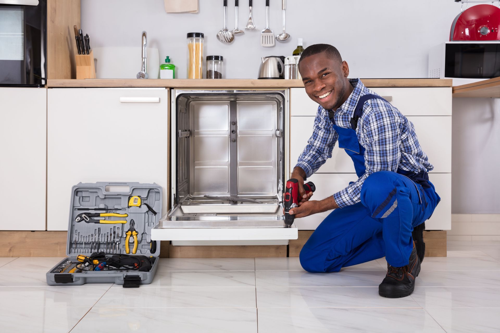 A person in blue jumpsuit repairs a dishwasher in a kitchen, smiling and holding a power drill; toolbox nearby.