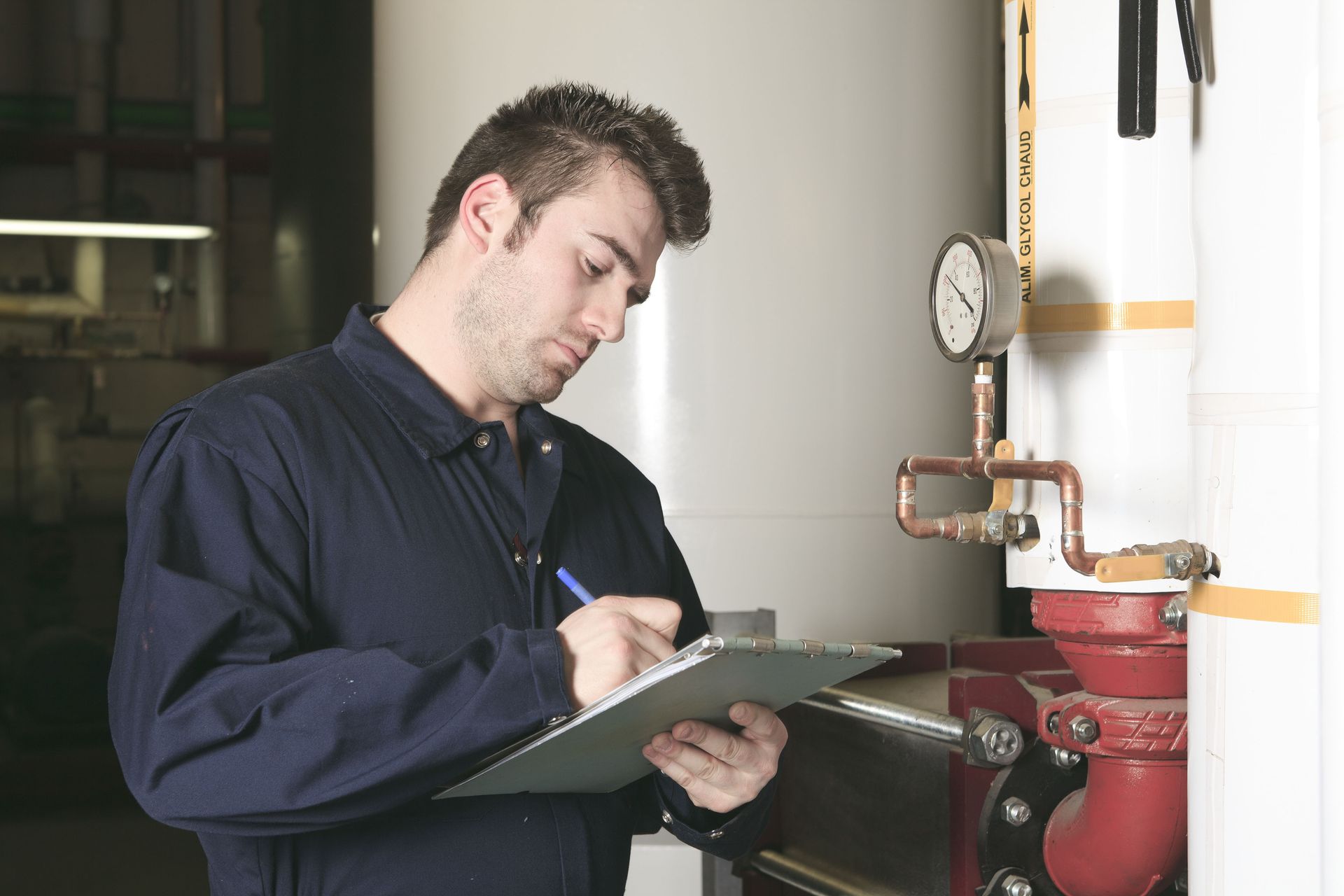 Man in blue jumpsuit, writing on a clipboard, inspecting machinery with gauges and pipes.