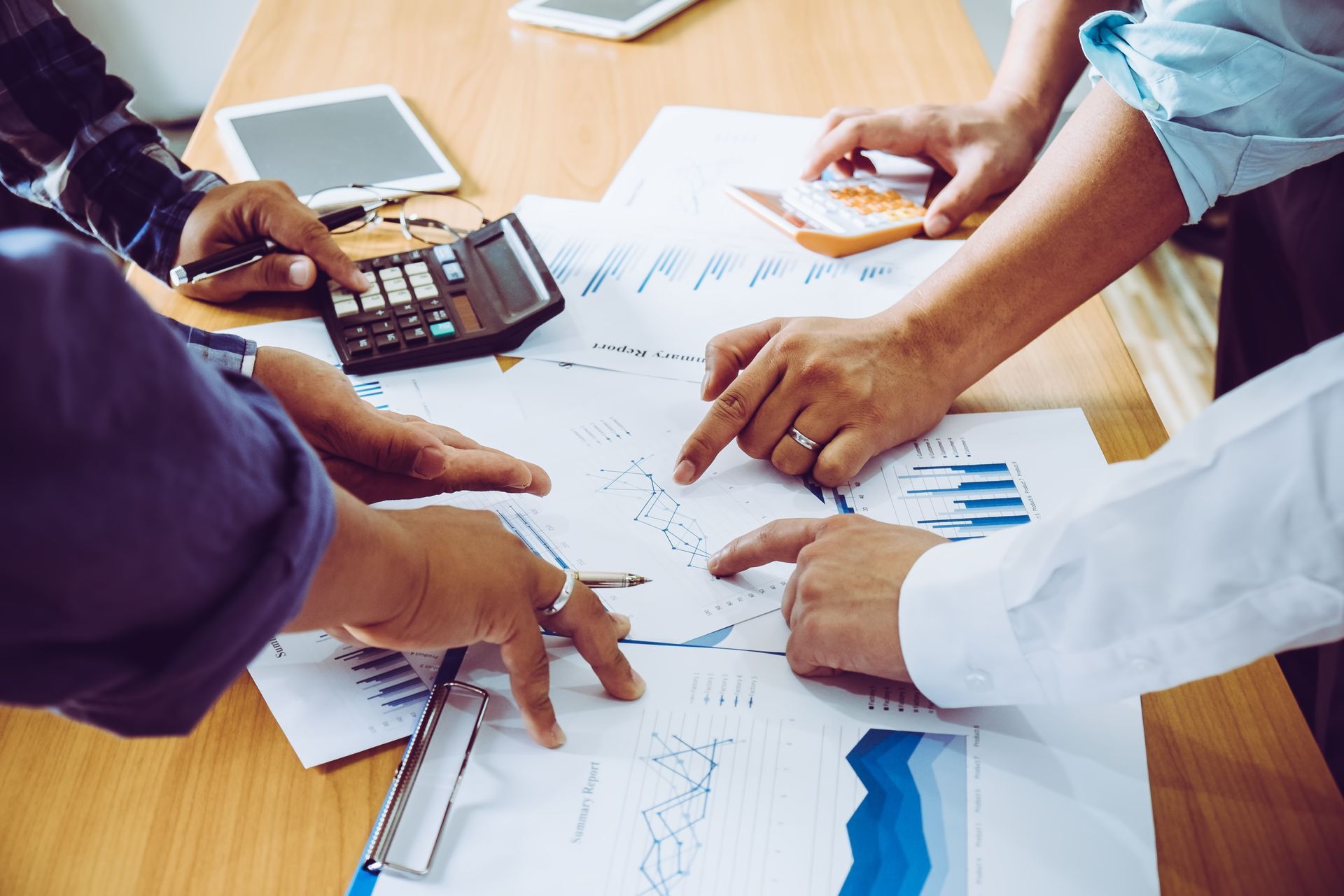 Hands pointing at financial reports on a table during a meeting, with a calculator and tablet.