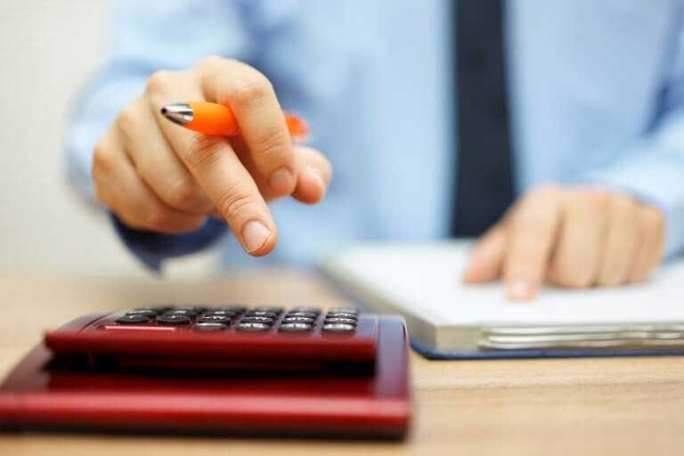 Person pointing at a calculator with an orange pen, working at a desk.