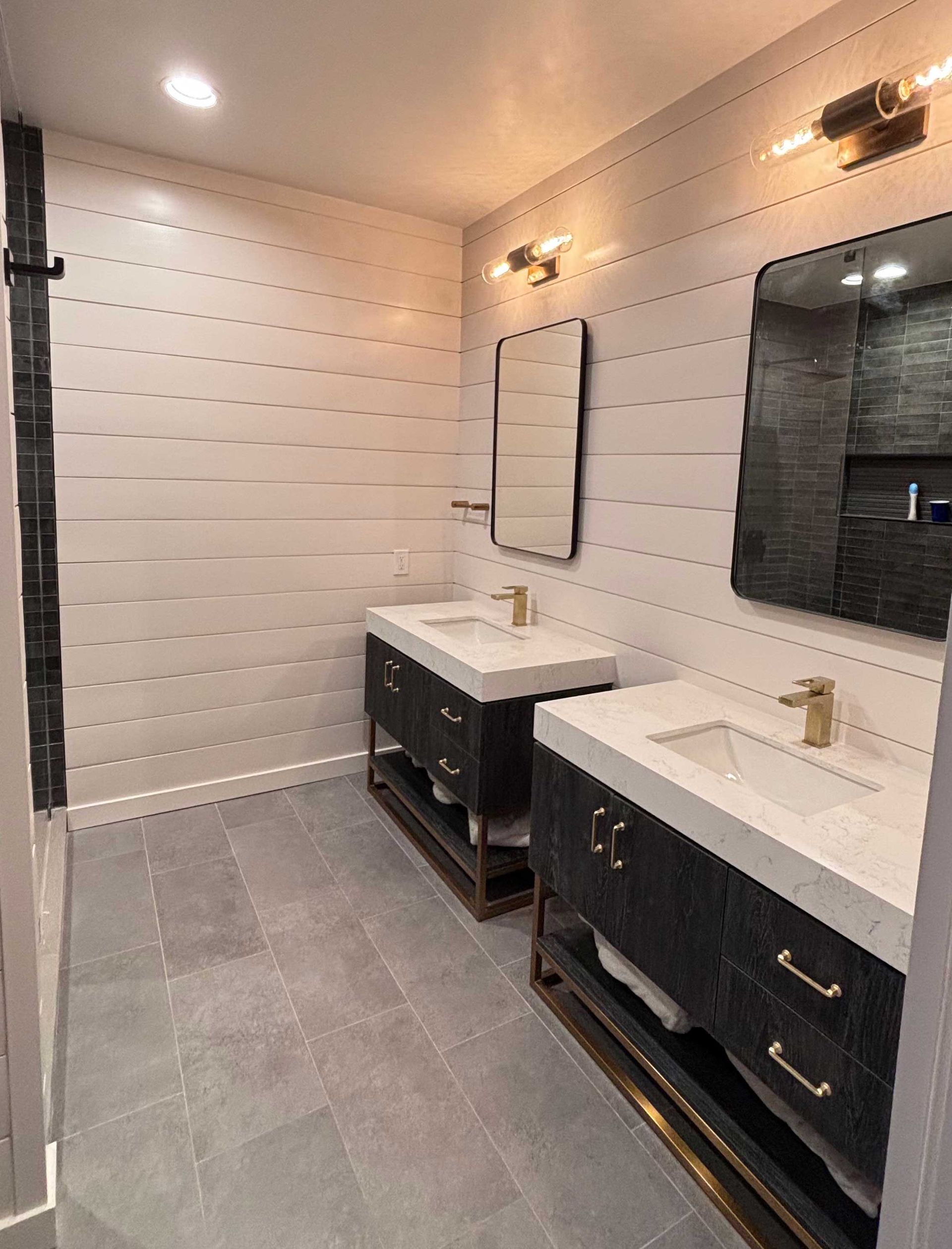 Bathroom with two sinks, white shiplap walls, gray tile floor, dark cabinets, gold fixtures.