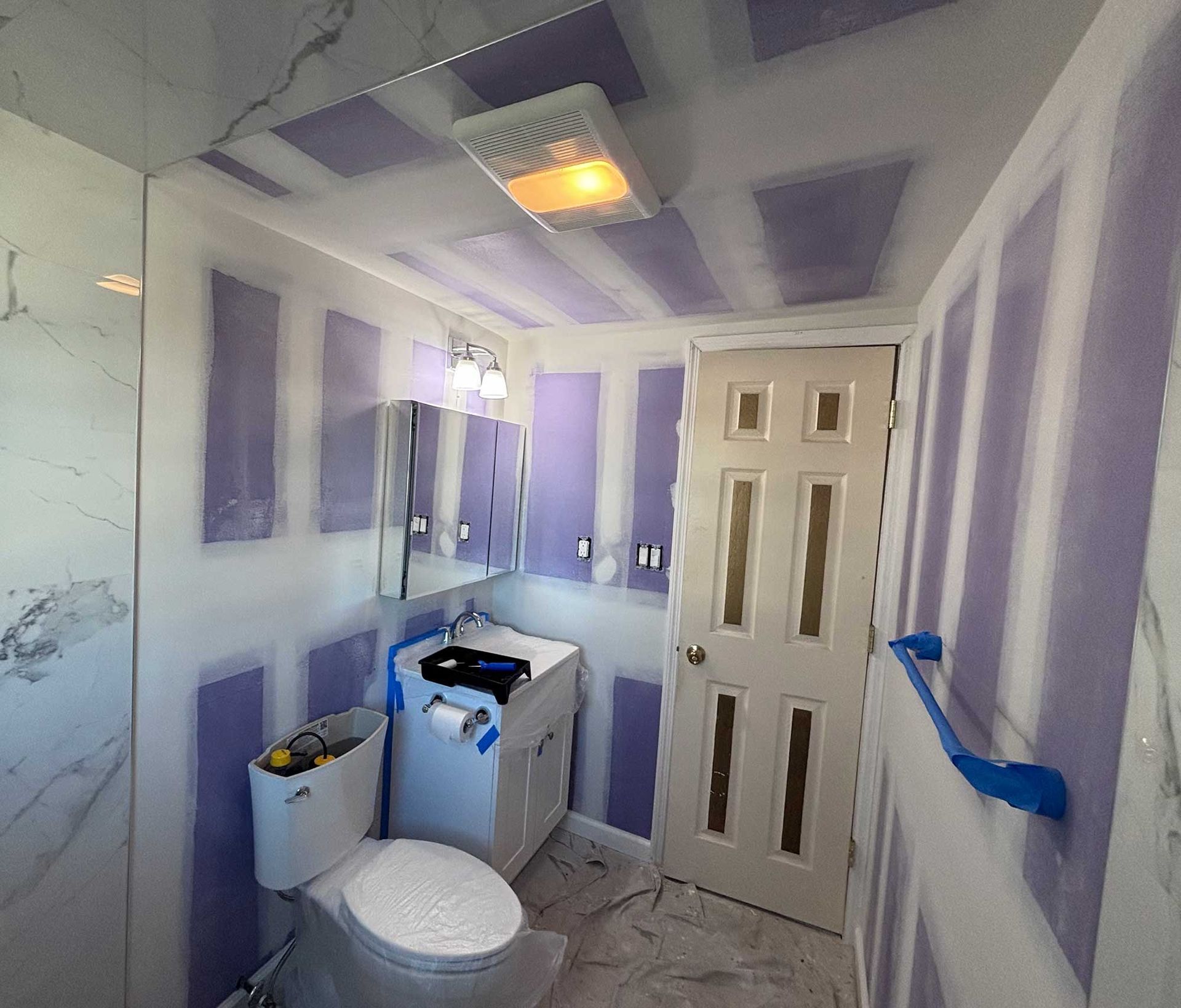 Bathroom under renovation, drywall installed. Light purple drywall, white toilet, vanity, and door.