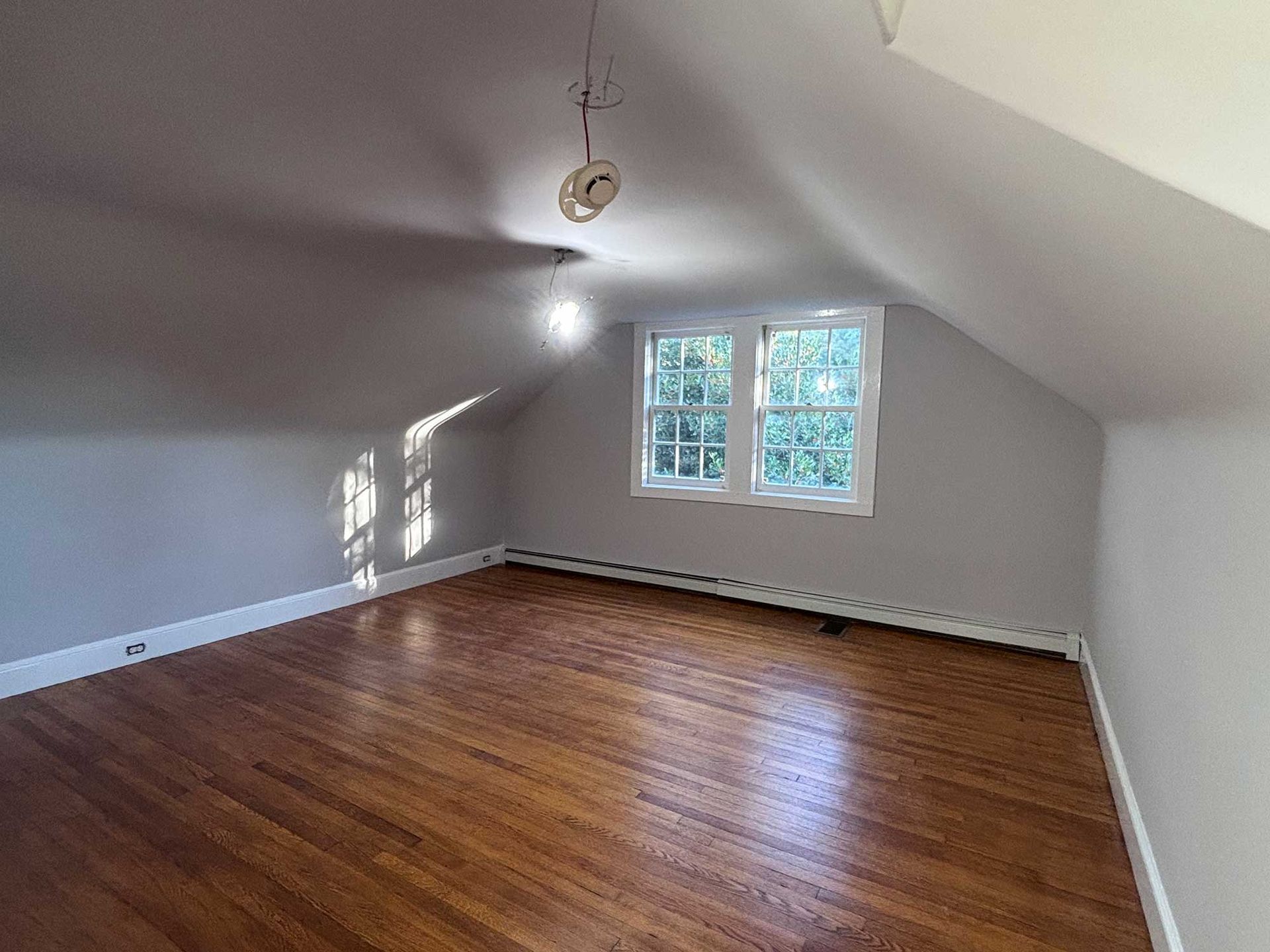 Empty attic room with hardwood floors, two windows, and slanted walls painted light gray.