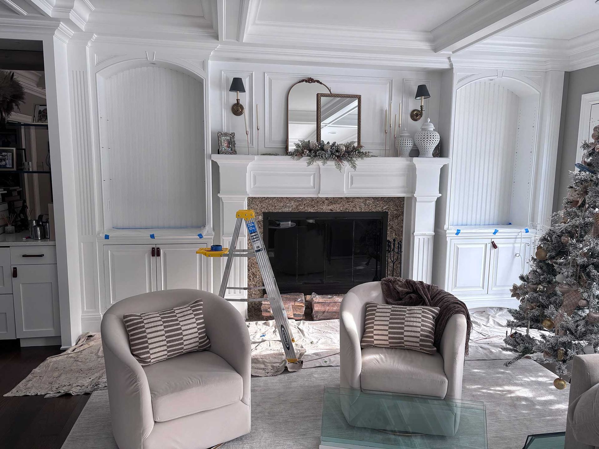 Living room with white fireplace, built-ins, and two armchairs. A ladder and Christmas tree are present.