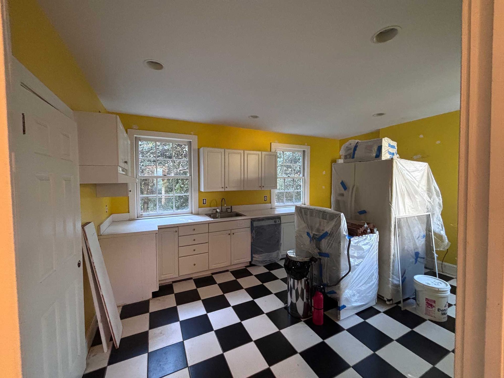 Kitchen with yellow walls, white cabinets, black and white checkered floor, covered appliances.