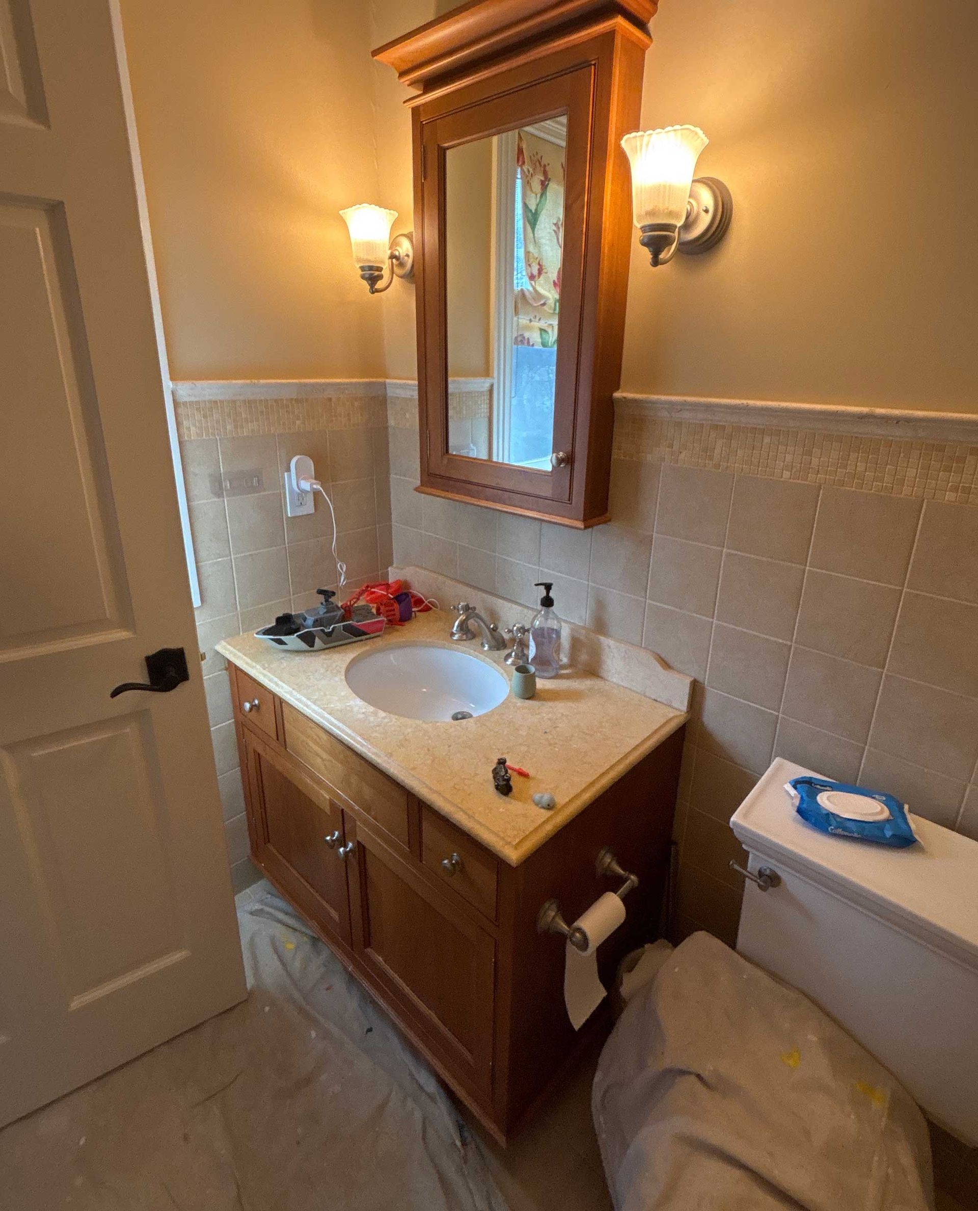Bathroom with wooden vanity, mirror, and toilet. Beige tiles on walls, two sconces above the vanity.