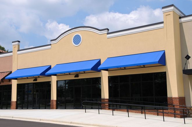 Exterior of a vacant commercial building with blue awnings over large windows, beige walls, and a ramp.
