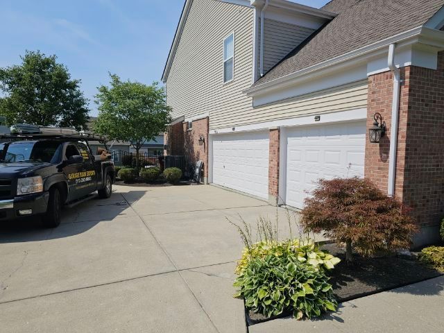 Suburban house driveway with two white garage doors, parked black SUV, and a flower bed by the entrance