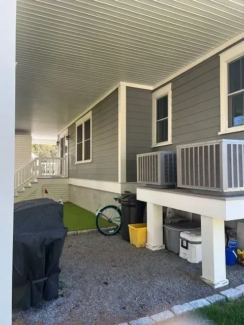 Covered side porch beside a gray house with a bike, grill, and storage bins on gravel.