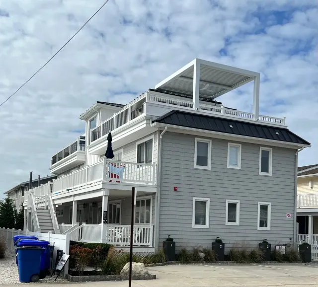 Gray multi-story house with white trim, porches, and rooftop deck under a partly cloudy sky