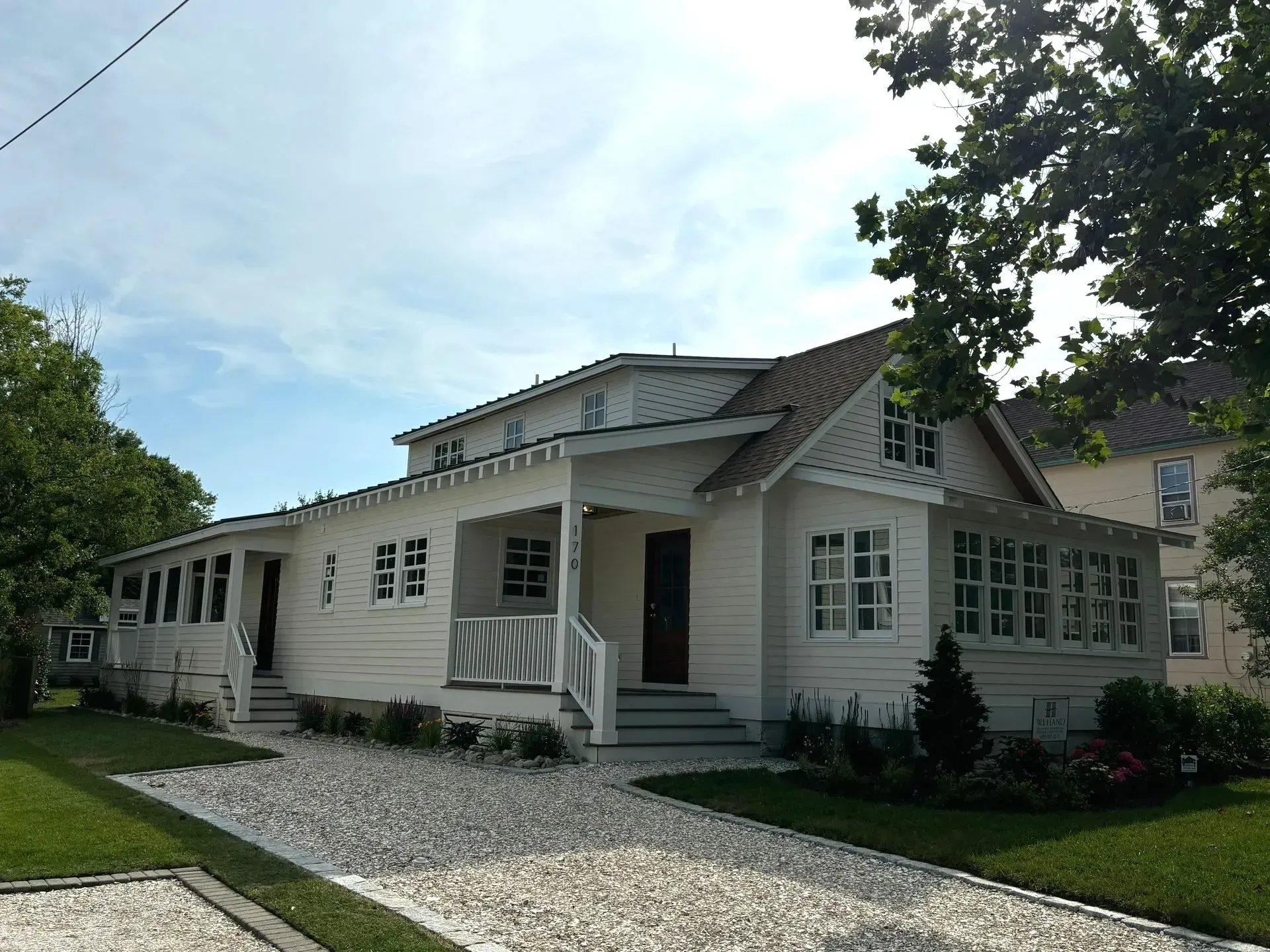 White single-story house with gray roof, front porch, and gravel path, surrounded by green lawn and trees