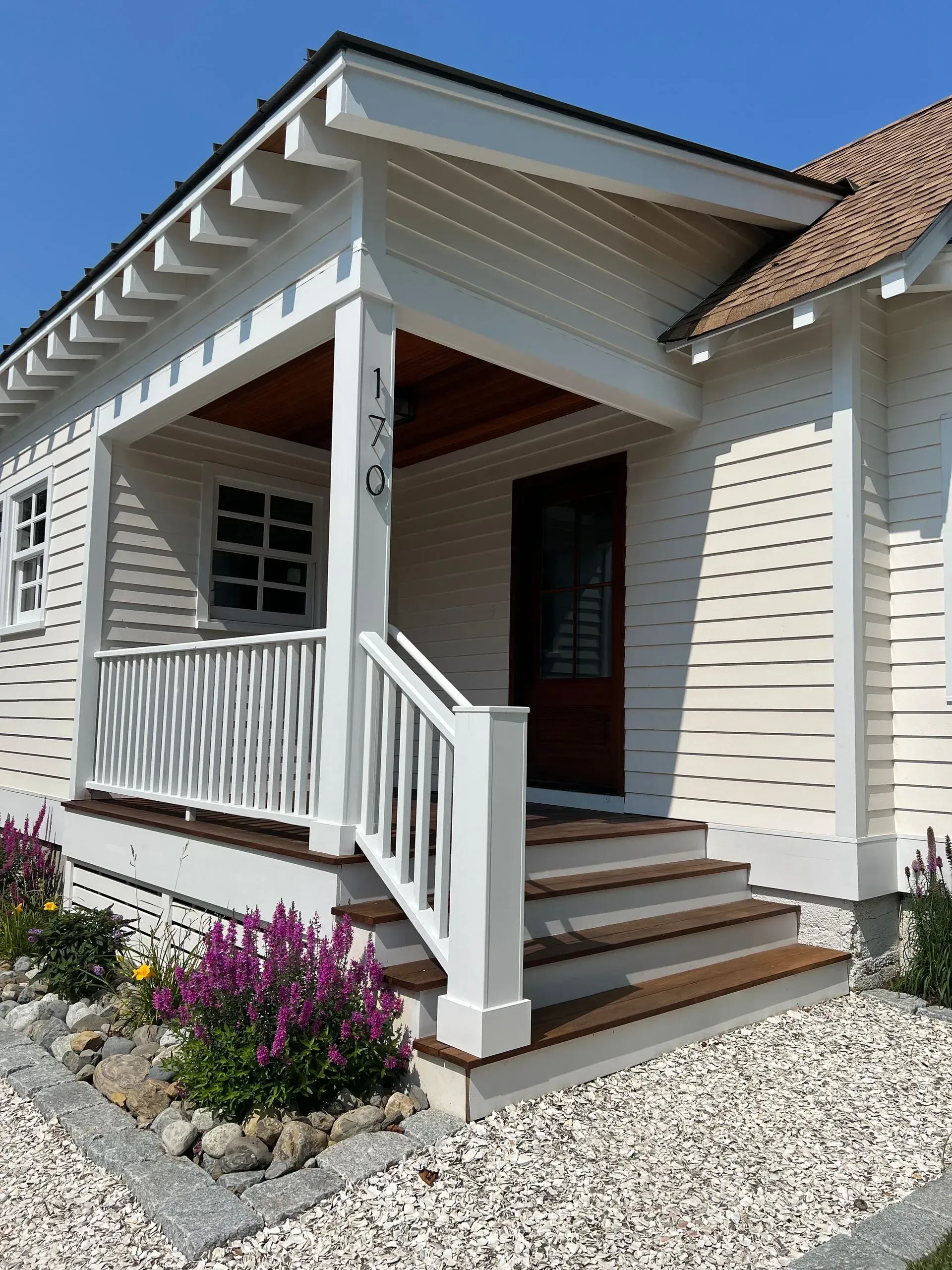 Front porch entrance with white railings, steps, and purple flowers beside a light-colored house.