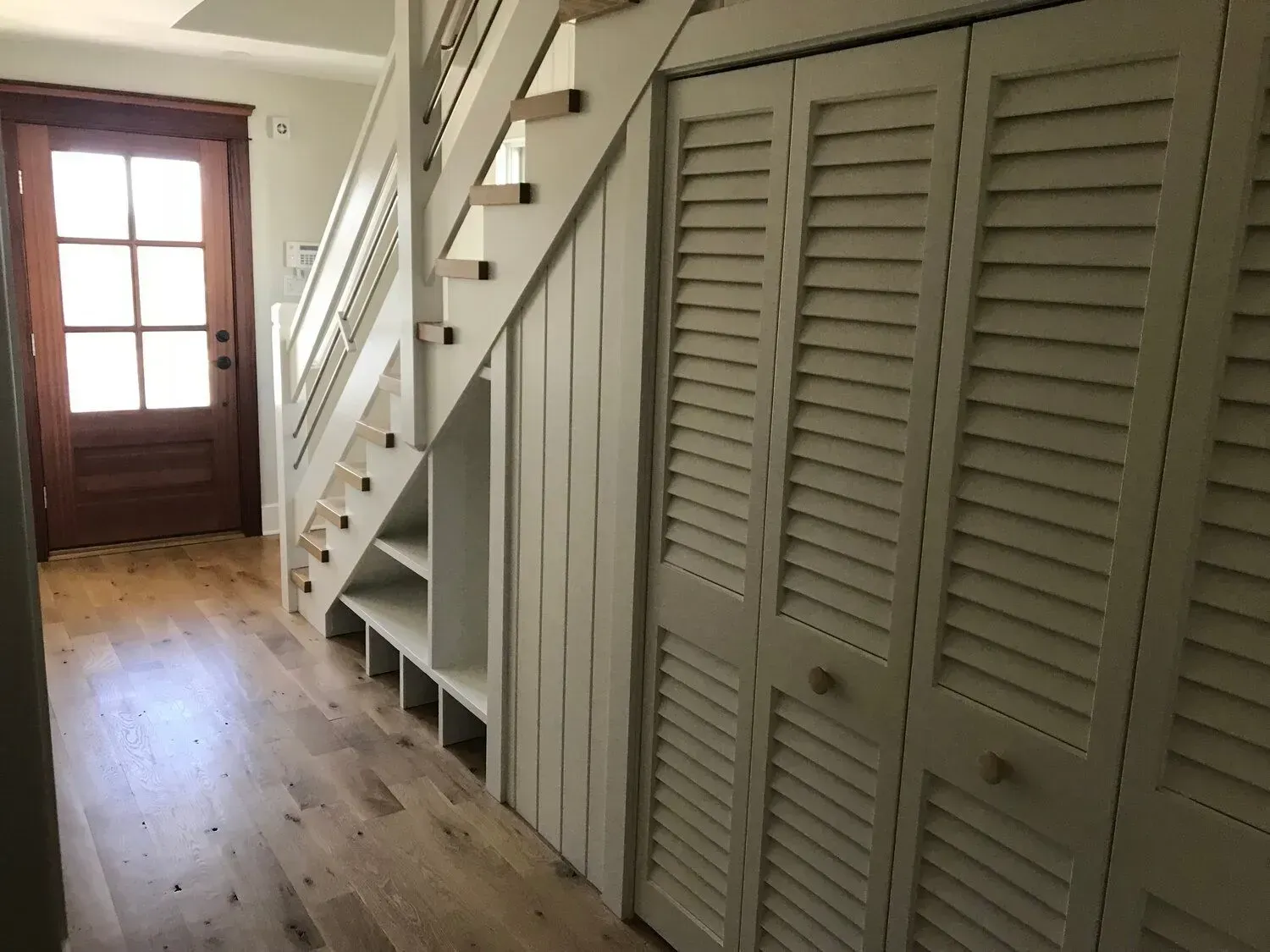 Bright hallway with a wooden floor, staircase, and white louvered closet doors near a front door.