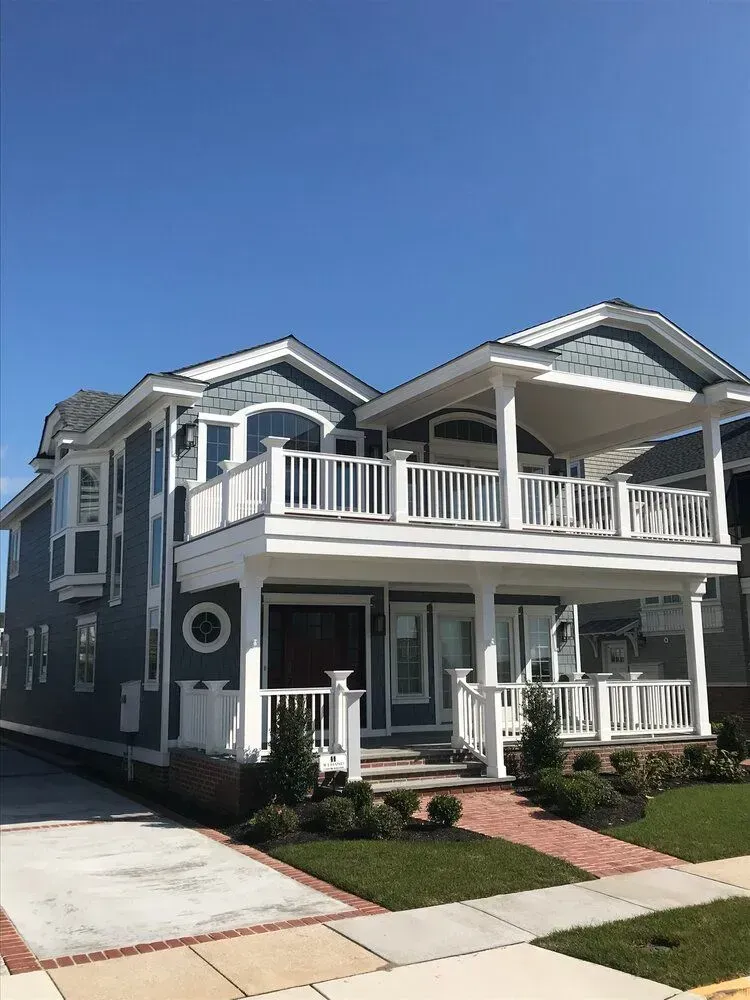 Two-story gray house with white trim, a covered porch, balcony, and driveway under a clear blue sky
