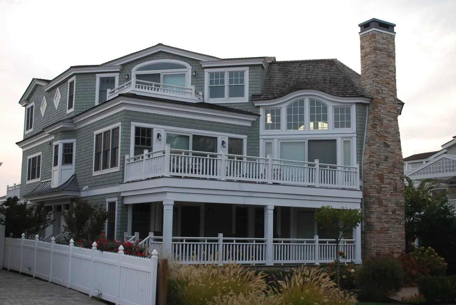 Large gray seaside house with white trim, wraparound porches, and a stone chimney at dusk