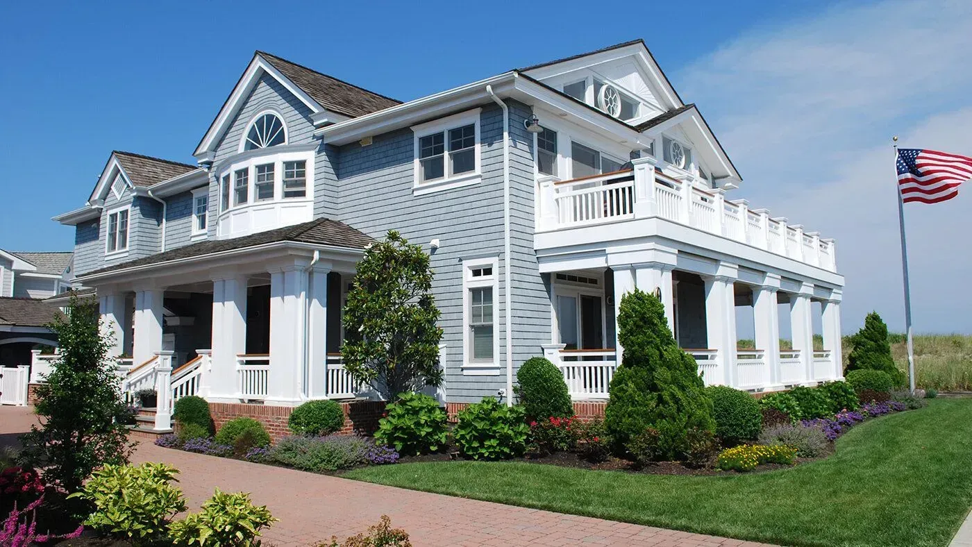Gray coastal-style house with white trim, porches, manicured landscaping, and an American flag