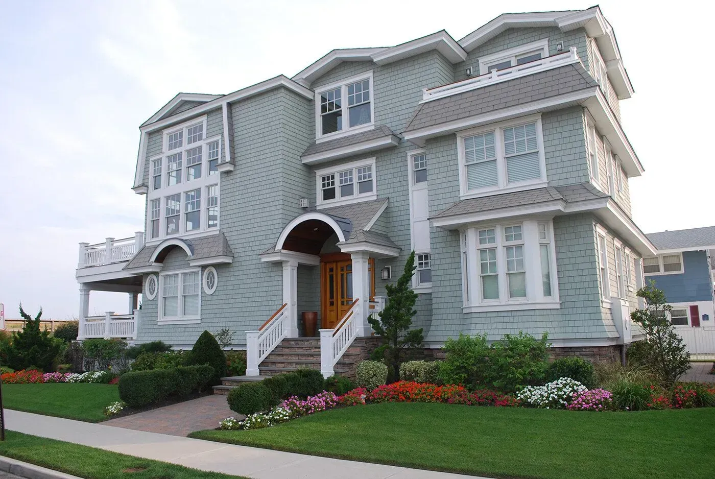 Large gray Victorian-style house with white trim, front steps, and manicured lawn and flower beds