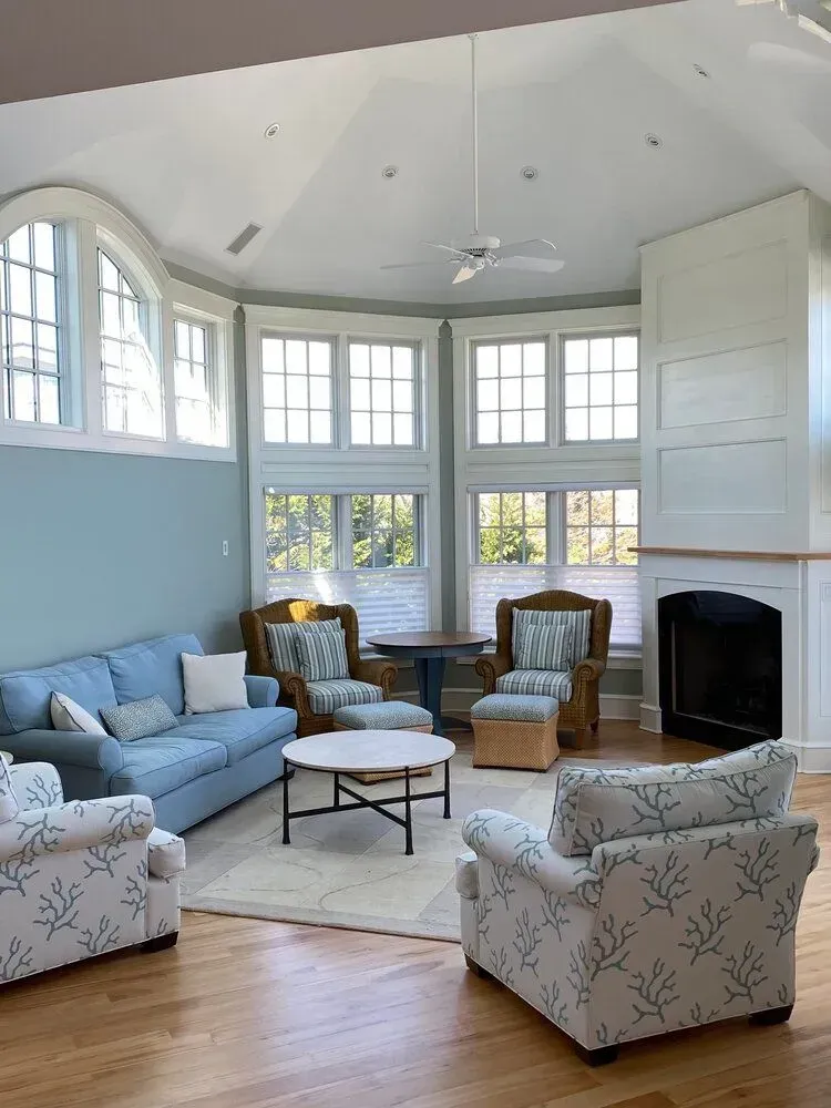 Bright living room with blue walls, large windows, fireplace, and patterned armchairs around a round table