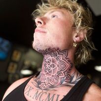 Man with neck tattoo of a geometric mandala and chest tattoo, looking up with a neutral expression.