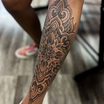 Detailed, gray ornamental tattoo on a person's lower leg. Person's foot in pink shoe is visible in the background.
