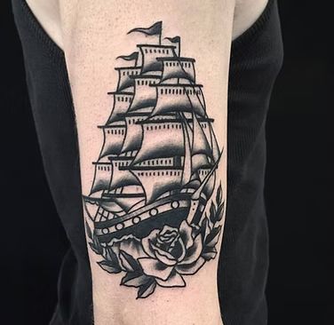 Black and white tattoo of a tall sailing ship, rose, and leaves on a person's upper arm.