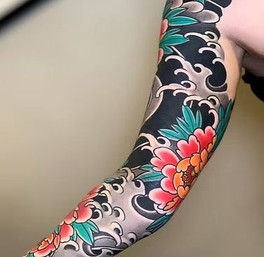 Full sleeve tattoo of red and orange peonies, green leaves, and black waves on an arm.