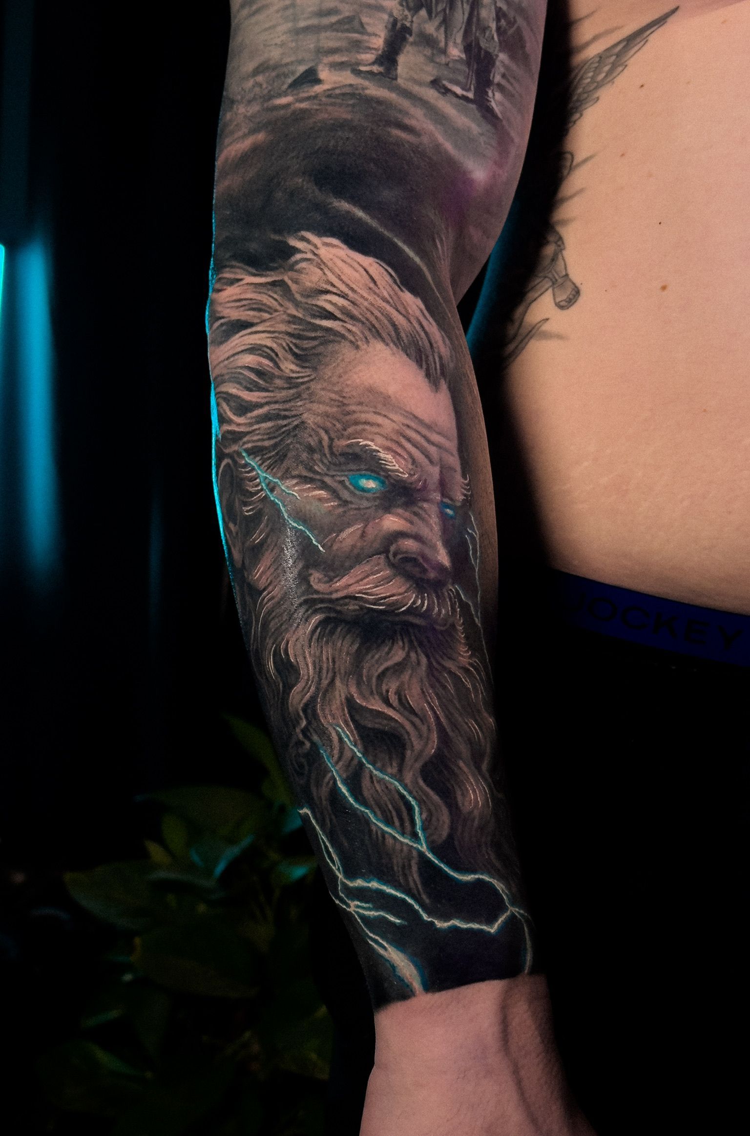 Realistic tattoo of a bearded man with glowing blue eyes, surrounded by lightning on an arm.