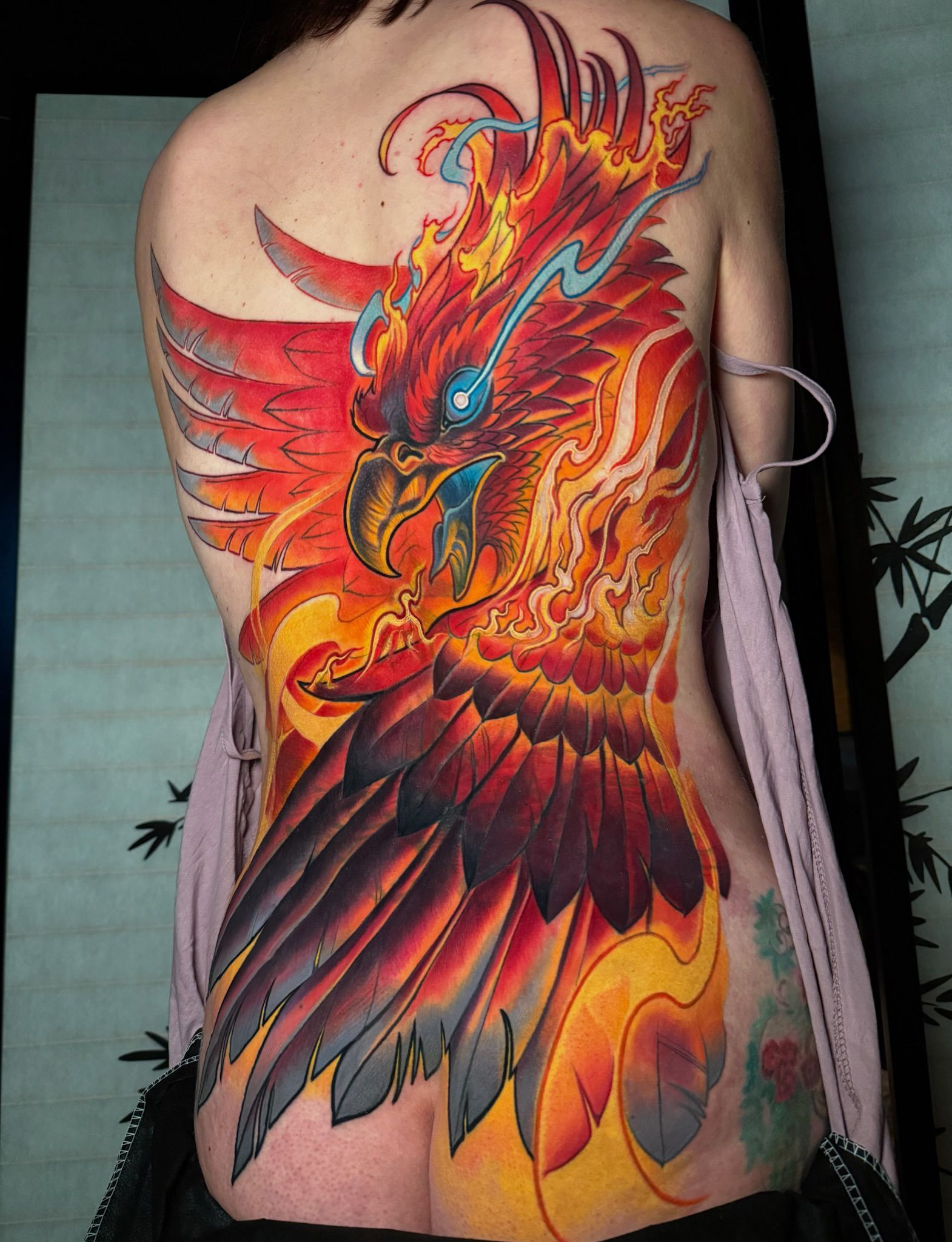 Vibrant phoenix tattoo spanning a person's back, in red, orange, and yellow with a blue eye.