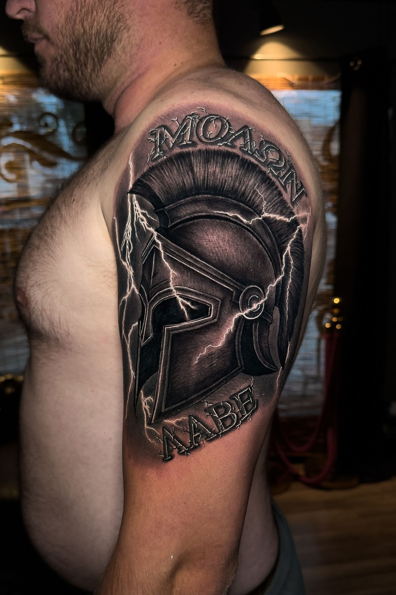 Tattoo of a Spartan helmet with lightning on a man's upper arm. The words 