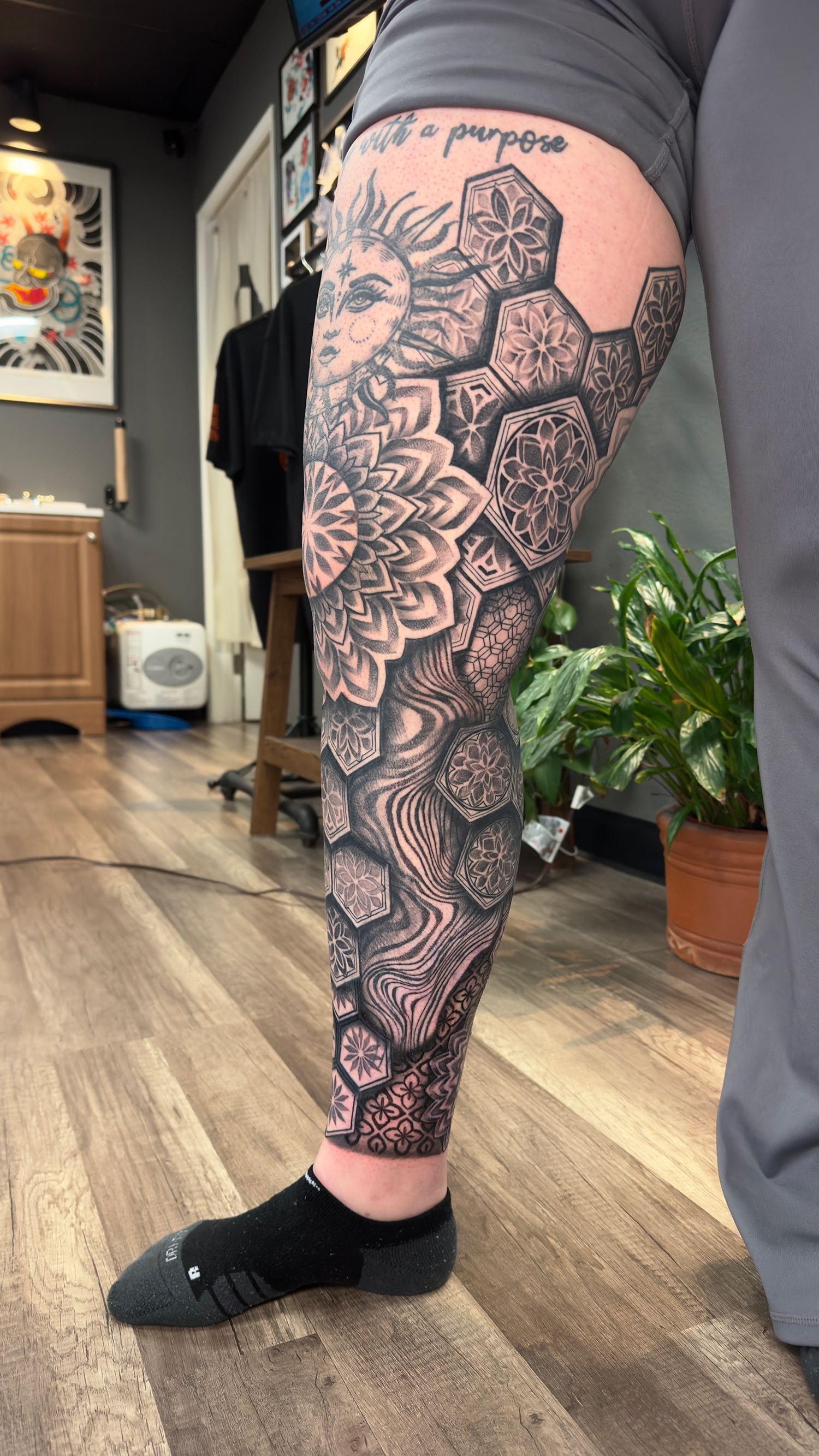 Leg with intricate black tattoo of sun, mandala, and honeycomb design. In a tattoo parlor setting.