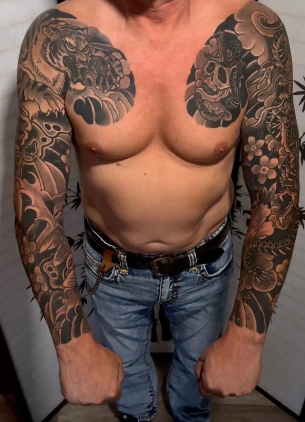 Man with full sleeve tattoos, wearing jeans, stands with fists clenched.