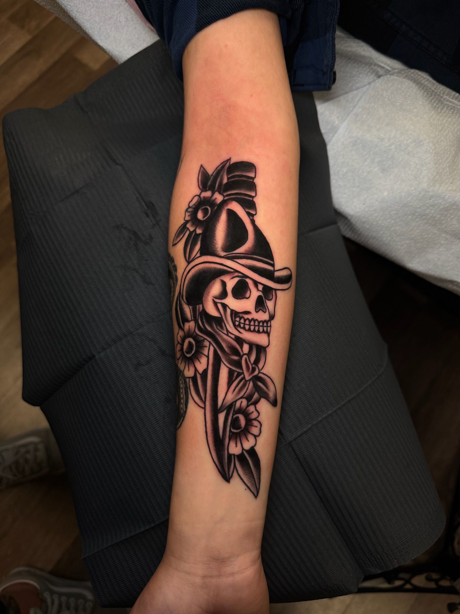 Tattoo of a skull wearing a hat and holding a dagger, surrounded by flowers on an arm.