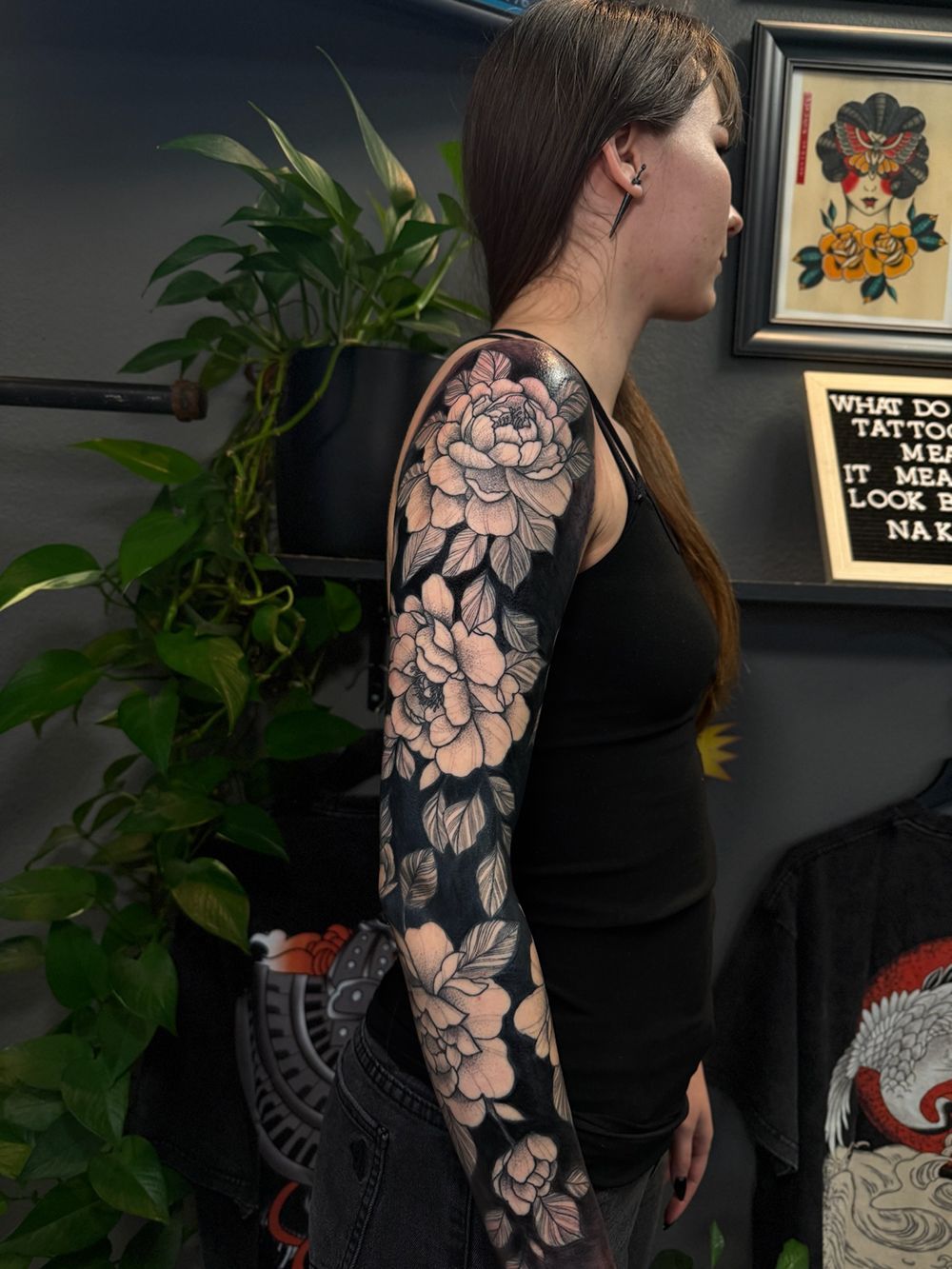 Woman with full sleeve floral tattoo stands in shop. Dark shirt, flowers in black and white.