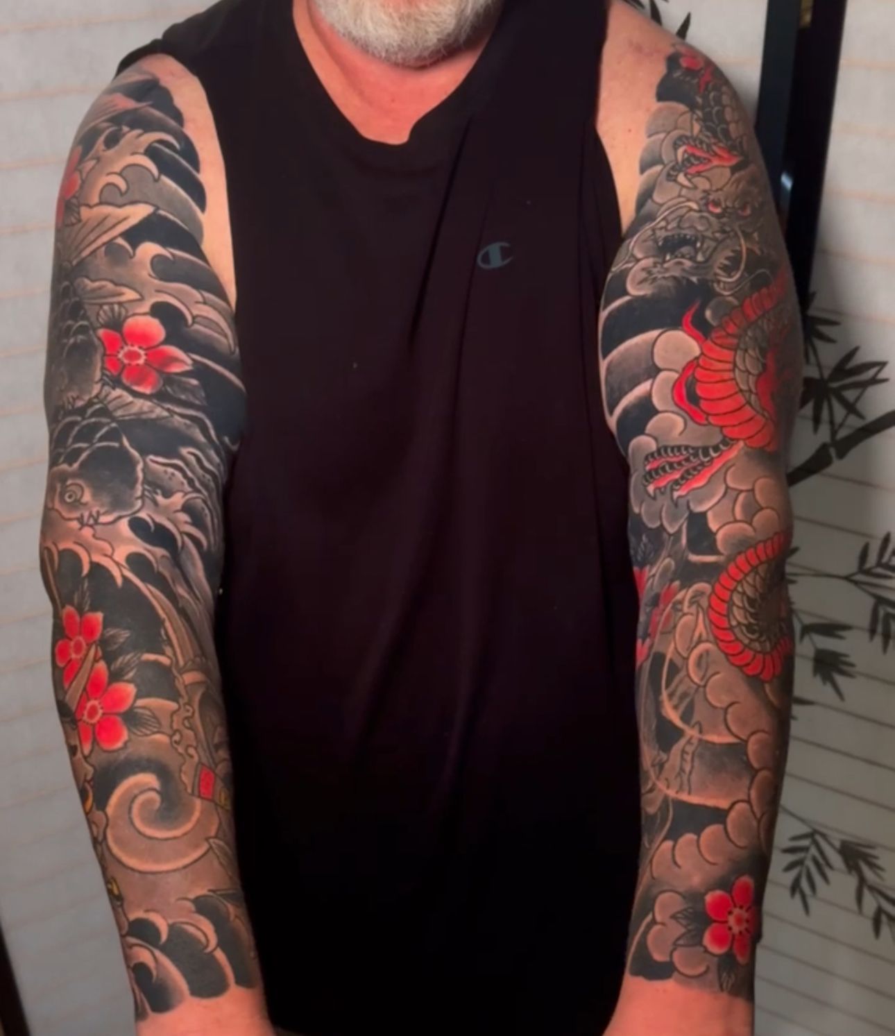 Man with full sleeve tattoos featuring a dragon, flowers, and waves in red, gray, and black.