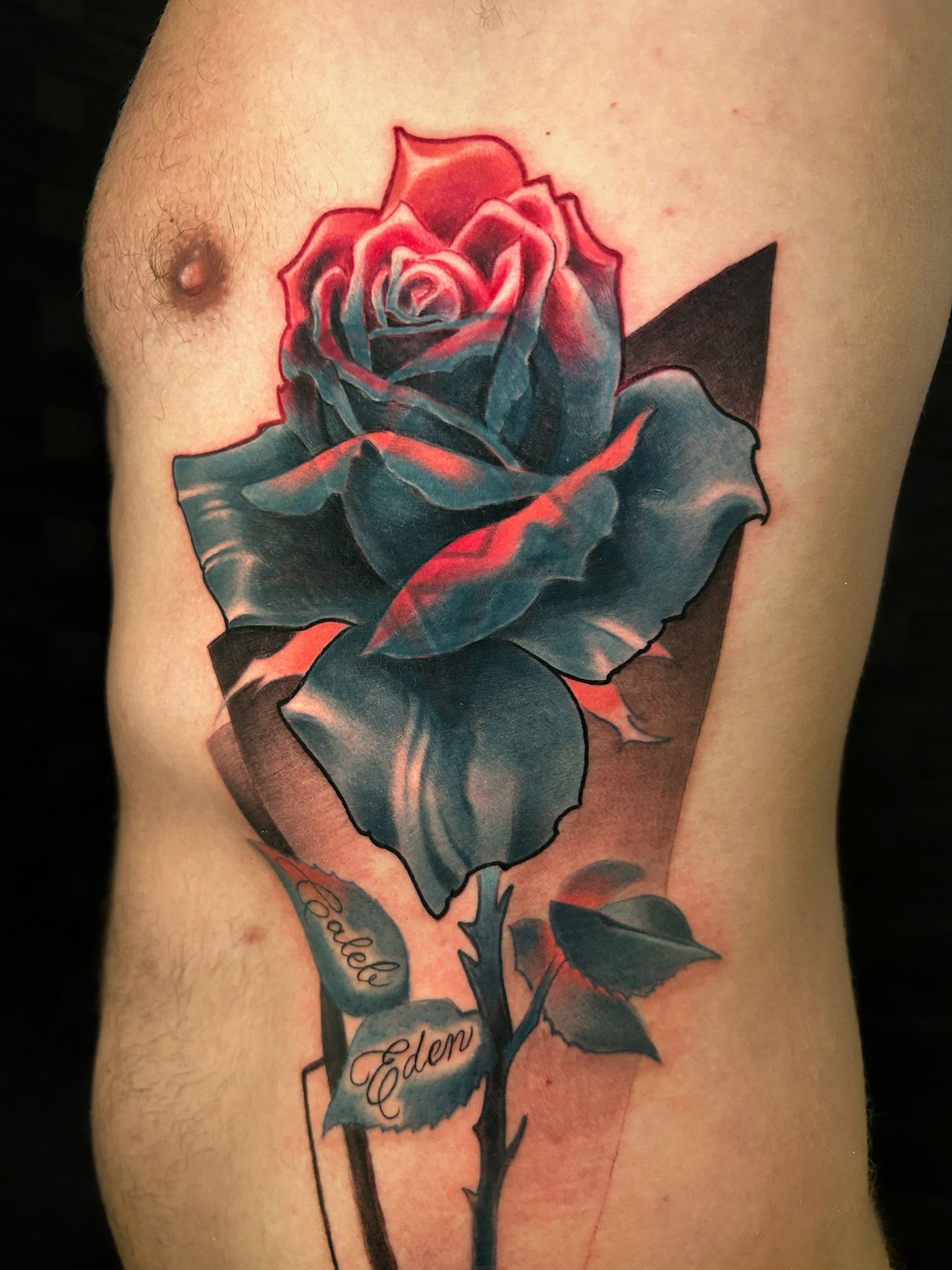 Tattoo of a rose on a torso. Teal and red flower with black geometric background.