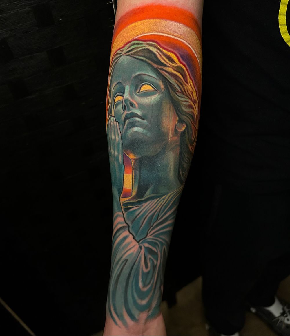 Tattoo of a serene statue with glowing eyes, set against an orange and yellow halo on the forearm.